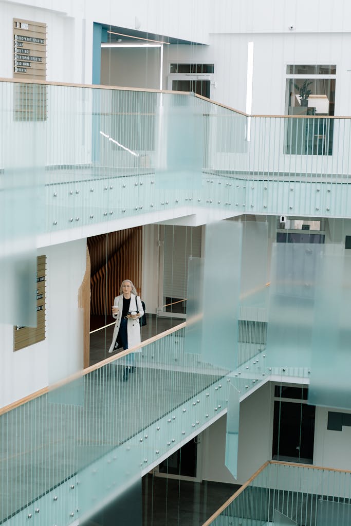 Startseite Kunden Woman walking through a bright, modern office corridor with glass railings, holding coffee and documents.
